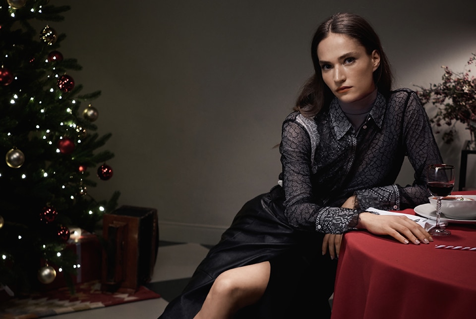 Junge Frau sitzt am Tisch, neben ihr steht ein geschmückter Weihnachtsbaum für die Luisa Cerano Weihnachtskampagne