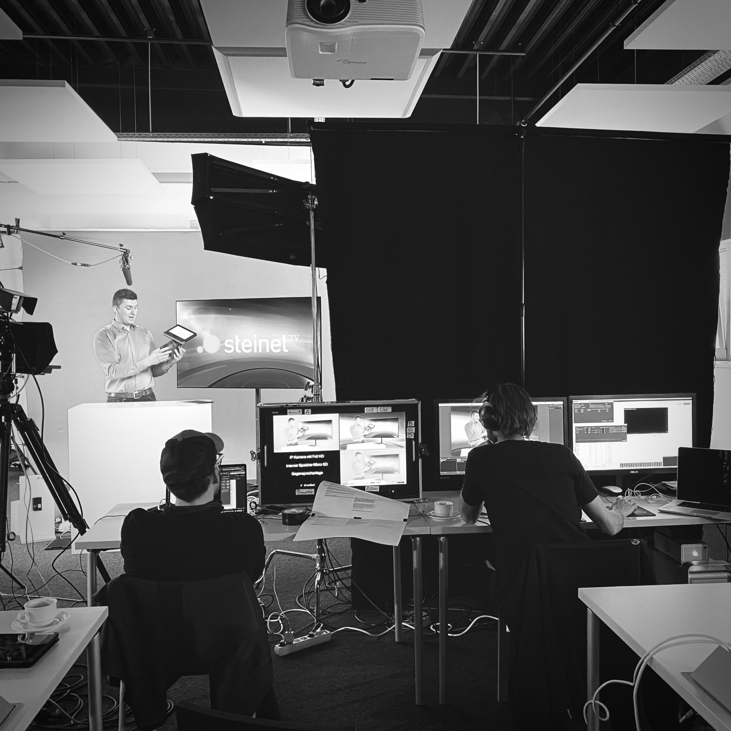 Ein Mann steht vor der Kamera und zwei Männer sitzen hinter Bildschirmen für die Produktion des Steinel TV Drehs